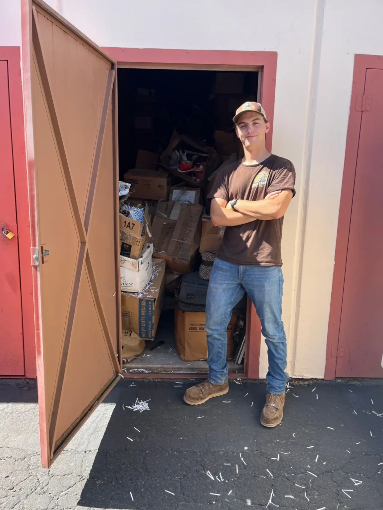 Junk removal specialist standing beside an open storage unit full of cardboard boxes and unwanted household items.